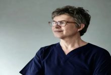 Fotoğrafta kısa gri saçlı, gözlüklü bir adam sade bir arka plan önünde yan tarafa doğru bakarken görülüyor. Lacivert sağlık kıyafeti giyen adam sakin ve kendinden emin bir ifadeyle poz veriyor.