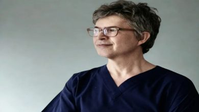 Fotoğrafta kısa gri saçlı, gözlüklü bir adam sade bir arka plan önünde yan tarafa doğru bakarken görülüyor. Lacivert sağlık kıyafeti giyen adam sakin ve kendinden emin bir ifadeyle poz veriyor.