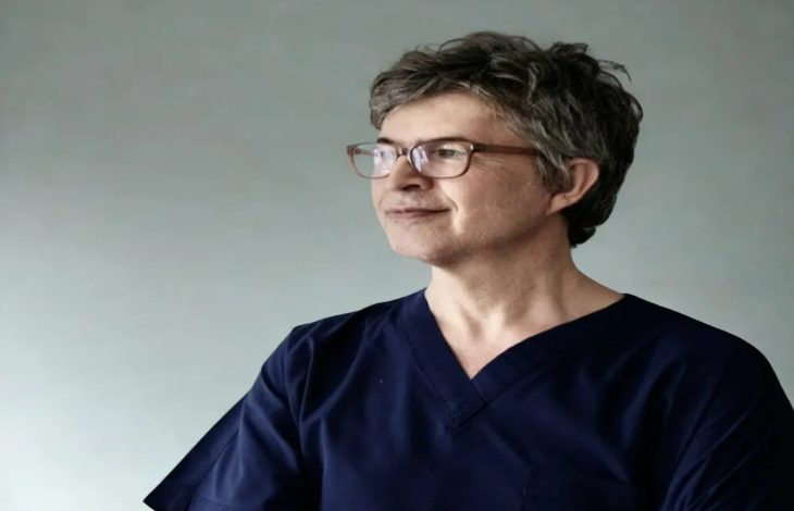 Fotoğrafta kısa gri saçlı, gözlüklü bir adam sade bir arka plan önünde yan tarafa doğru bakarken görülüyor. Lacivert sağlık kıyafeti giyen adam sakin ve kendinden emin bir ifadeyle poz veriyor.