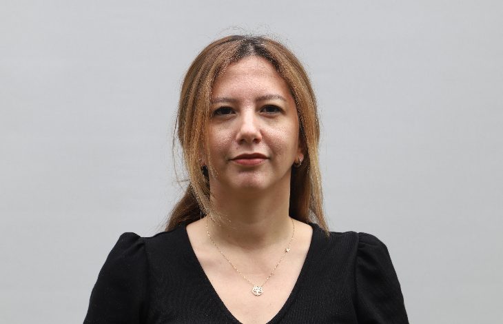 Düz, açık renkli bir arka plan önünde çekilmiş bir portre. Omuz hizasında kadraja alınmış bir kadın doğrudan kameraya bakıyor. Siyah bir bluz giymiş ve ince bir kolye takıyor. Yüz ifadesi sakin ve ciddi; fotoğraf kurumsal ya da haber amaçlı bir çekim izlenimi veriyor.