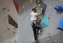 Görselde, bir sporcu yapay tırmanış duvarında yukarı doğru ilerliyor. Duvar üzerinde farklı renklerde tutamaklar bulunuyor ve sporcu emniyet ipine bağlı durumda. Odak, tırmanış sırasında sporcuların güç ve denge gerektiren hareketlerine verilmiş. Arka planda Fethiye’de düzenlenen bir para tırmanış antrenmanının atmosferi yansıyor.