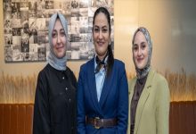 Kapalı bir mekânda yan yana duran üç kadın kameraya gülümseyerek poz veriyor. İkisi başörtülü, biri resmi bir takım elbise giymiş. Arka planda fotoğraflardan oluşan bir pano ve sade bir iç mekân dekoru görülüyor.