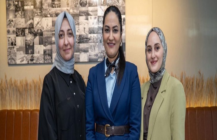 Kapalı bir mekânda yan yana duran üç kadın kameraya gülümseyerek poz veriyor. İkisi başörtülü, biri resmi bir takım elbise giymiş. Arka planda fotoğraflardan oluşan bir pano ve sade bir iç mekân dekoru görülüyor.