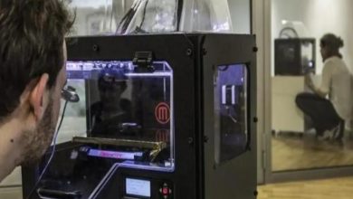 Bir laboratuvar ortamında, bir kişi masa üzerindeki 3D yazıcıyı yakından izliyor. Cihazın içinde üretim süreci devam ederken, arka planda cam bölmenin arkasında başka bir kişinin benzer bir makineyle çalıştığı görülüyor. Ortam teknik ve araştırma odaklı bir çalışma alanını andırıyor.