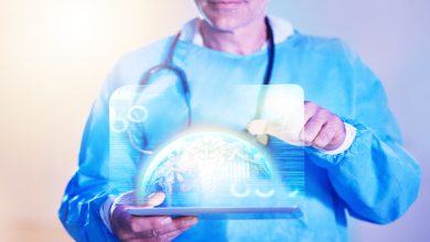 Fotoğrafta sağlık çalışanı kıyafeti giymiş bir kişinin, elinde tablet tutarak dijital bir sağlık arayüzünü kullandığı görülüyor. Kişi sanal gerçeklik gözlüğü takıyor ve tablet üzerinde hologram şeklinde dünya küresi ile tıbbi veriler yer alıyor. Görsel, sağlık teknolojileri ve dijital tıp temasını yansıtıyor.