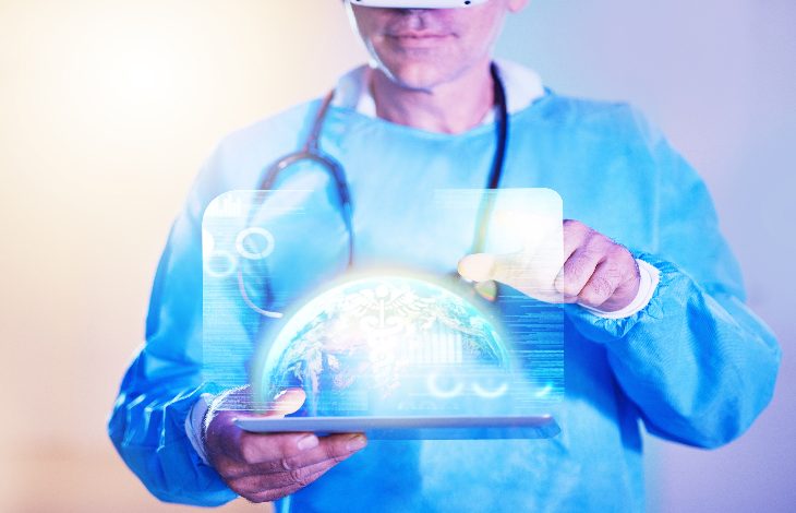 Fotoğrafta sağlık çalışanı kıyafeti giymiş bir kişinin, elinde tablet tutarak dijital bir sağlık arayüzünü kullandığı görülüyor. Kişi sanal gerçeklik gözlüğü takıyor ve tablet üzerinde hologram şeklinde dünya küresi ile tıbbi veriler yer alıyor. Görsel, sağlık teknolojileri ve dijital tıp temasını yansıtıyor.