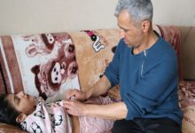 Ev ortamında çekilmiş görüntüde, orta yaşlı bir adam koltukta uzanan küçük bir çocuğun kıyafetini düzeltirken ona özenle bakıyor. Sahne, bakım ve sorumluluğun sessiz ama güçlü bir anını yansıtıyor.