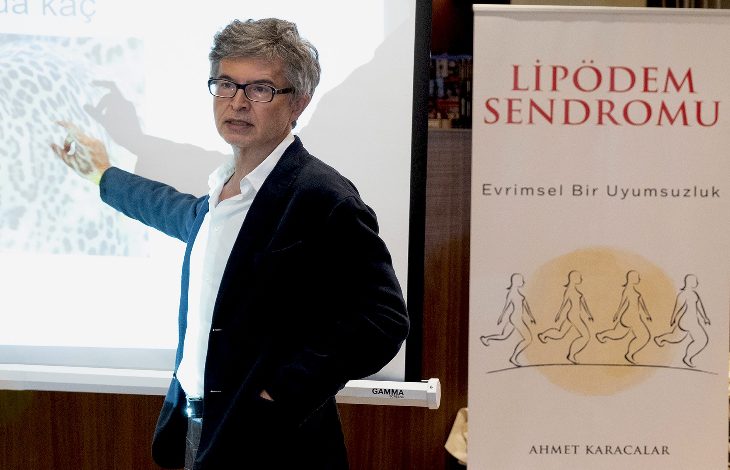 Bir sunum sırasında takım elbiseli bir uzman, projeksiyon ekranını işaret ederek anlatım yapıyor. Yanında “Lipödem Sendromu” başlıklı bir afiş yer alıyor; sahne, tıbbi bir bilgilendirme ya da konferans ortamını yansıtıyor.