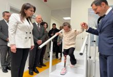 Fotoğrafta protez bacak kullanan küçük bir kız çocuğu merdivenlerden inerken bir erkek tarafından elinden tutularak destekleniyor. Çevrede bulunan resmi kıyafetli kişiler çocuğu dikkatle izlerken, kız çocuğu protez bacağıyla adım atarak yürümeyi deniyor. Arka planda bir sağlık ya da rehabilitasyon merkezinin koridoru görülüyor.