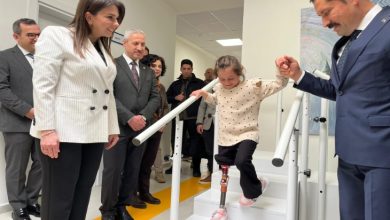 Fotoğrafta protez bacak kullanan küçük bir kız çocuğu merdivenlerden inerken bir erkek tarafından elinden tutularak destekleniyor. Çevrede bulunan resmi kıyafetli kişiler çocuğu dikkatle izlerken, kız çocuğu protez bacağıyla adım atarak yürümeyi deniyor. Arka planda bir sağlık ya da rehabilitasyon merkezinin koridoru görülüyor.
