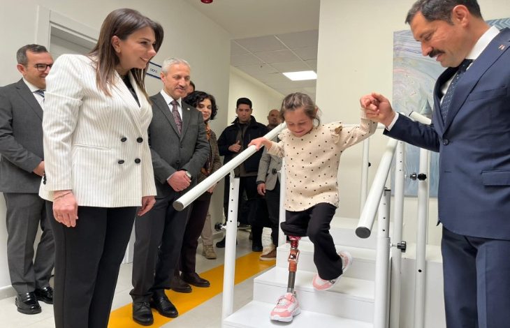Fotoğrafta protez bacak kullanan küçük bir kız çocuğu merdivenlerden inerken bir erkek tarafından elinden tutularak destekleniyor. Çevrede bulunan resmi kıyafetli kişiler çocuğu dikkatle izlerken, kız çocuğu protez bacağıyla adım atarak yürümeyi deniyor. Arka planda bir sağlık ya da rehabilitasyon merkezinin koridoru görülüyor.