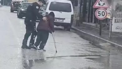Yağmurlu bir günde çekilen görüntüde, bir polis memuru bastonla yürüyen engelli bir bireyin koluna destek olarak onu yolun karşısına geçirmeye yardımcı oluyor. Islak yolda trafik akışı görülürken, polis memurunun dikkatli şekilde eşlik ettiği an objektife yansımış.