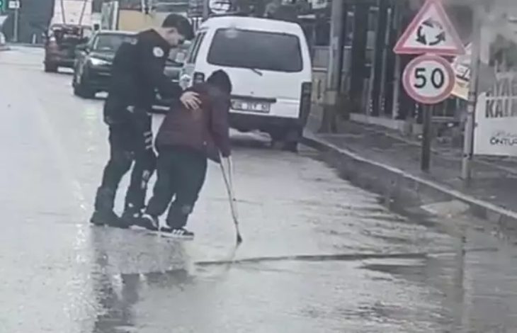 Yağmurlu bir günde çekilen görüntüde, bir polis memuru bastonla yürüyen engelli bir bireyin koluna destek olarak onu yolun karşısına geçirmeye yardımcı oluyor. Islak yolda trafik akışı görülürken, polis memurunun dikkatli şekilde eşlik ettiği an objektife yansımış.