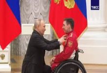 Resmi bir törende, tekerlekli sandalyedeki bir sporcuya Rusya Devlet Başkanı Vladimir Putin tarafından madalya takdim ediliyor. Arka planda bayraklar ve kürsü bulunuyor. Sahne, uluslararası bir başarıyı ve ödüllendirme anını yansıtıyor.