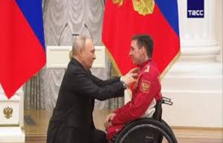 Resmi bir törende, tekerlekli sandalyedeki bir sporcuya Rusya Devlet Başkanı Vladimir Putin tarafından madalya takdim ediliyor. Arka planda bayraklar ve kürsü bulunuyor. Sahne, uluslararası bir başarıyı ve ödüllendirme anını yansıtıyor.