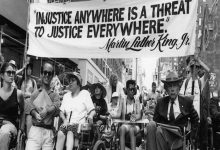 Şehrin merkezinde, yüksek binaların arasında ilerleyen bir grup engelli aktivist, tekerlekli sandalyeleriyle güçlü ve kararlı bir protesto yürüyüşü gerçekleştiriyor. Kalabalığın önünde taşınan pankartta Martin Luther King Jr.’ın şu sözü yer alıyor: “Injustice anywhere is a threat to justice everywhere.” (Herhangi bir yerdeki adaletsizlik, her yerdeki adalet için tehdittir.) Bu ifade yalnızca bir alıntı değil; eşitlik, erişilebilirlik ve hak temelli mücadelenin evrensel çağrısını simgeleyen güçlü bir hatırlatma niteliği taşıyor. Yürüyüş, dayanışmanın görünür hâle geldiği, adalet talebinin kamusal alanda kararlılıkla dile getirildiği bir hak savunuculuğu örneği olarak öne çıkıyor.