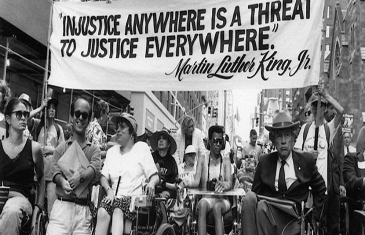 Şehrin merkezinde, yüksek binaların arasında ilerleyen bir grup engelli aktivist, tekerlekli sandalyeleriyle güçlü ve kararlı bir protesto yürüyüşü gerçekleştiriyor. Kalabalığın önünde taşınan pankartta Martin Luther King Jr.’ın şu sözü yer alıyor: “Injustice anywhere is a threat to justice everywhere.” (Herhangi bir yerdeki adaletsizlik, her yerdeki adalet için tehdittir.) Bu ifade yalnızca bir alıntı değil; eşitlik, erişilebilirlik ve hak temelli mücadelenin evrensel çağrısını simgeleyen güçlü bir hatırlatma niteliği taşıyor. Yürüyüş, dayanışmanın görünür hâle geldiği, adalet talebinin kamusal alanda kararlılıkla dile getirildiği bir hak savunuculuğu örneği olarak öne çıkıyor.