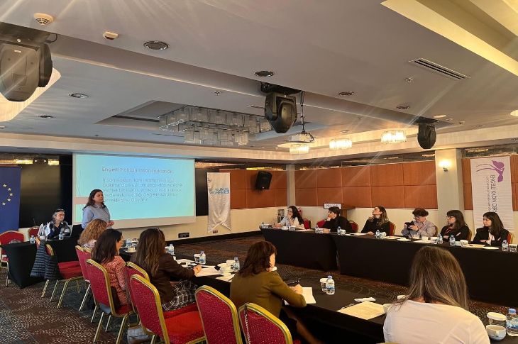 Bir otel konferans salonunda düzenlenen toplantıda, katılımcılar U şeklinde yerleştirilmiş masalarda oturuyor. Ön tarafta bir konuşmacı sunum yaparken projeksiyon ekranında “engelliliğe dayalı ayrımcılık” konulu bir slayt yer alıyor. Masalarda notlar ve su şişeleri bulunurken, katılımcılar dikkatle sunumu dinliyor. Solda Avrupa Birliği bayrağı, sağda ise UN Women afişi dikkat çekiyor.