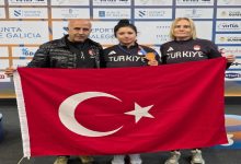 Görselde bir sporcu kazandığı bronz madalya ile Türkiye bayrağını iki kişiyle birlikte tutarak poz veriyor. Üç kişi “Türkiye” yazılı eşofmanlarla şampiyona alanında gururla fotoğraf çektiriyor. Arkada organizasyonun sponsor panosu yer alıyor.