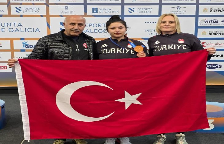 Görselde bir sporcu kazandığı bronz madalya ile Türkiye bayrağını iki kişiyle birlikte tutarak poz veriyor. Üç kişi “Türkiye” yazılı eşofmanlarla şampiyona alanında gururla fotoğraf çektiriyor. Arkada organizasyonun sponsor panosu yer alıyor.