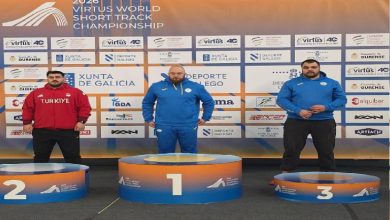 Görselde uluslararası bir atletizm şampiyonasının ödül töreni görülüyor. Üç sporcu podyumda yer alırken, ikinci sıradaki sporcu Türkiye eşofmanıyla kürsüde duruyor. Arkada yarışmanın sponsor ve organizasyon panosu bulunuyor.