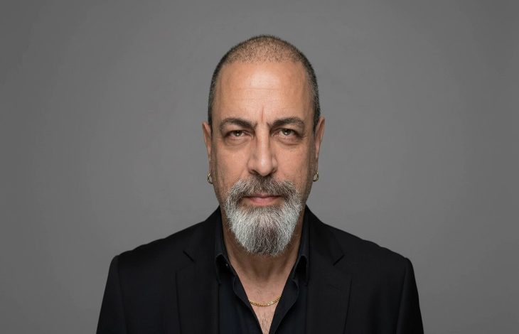 Gri arka plan önünde çekilmiş bir portre fotoğrafı görülüyor. Orta yaşlı bir erkek doğrudan kameraya bakıyor. Kısa kesilmiş saçları, belirgin kaşları ve gri-beyaz karışımı sakalı var. Siyah bir ceket ve koyu renk gömlek giymiş; kulağında küçük halka küpeler ve boynunda ince bir zincir bulunuyor. Yüz ifadesi ciddi ve sakin.