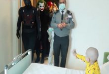 Bir hastane odasında, yatağın yanında duran küçük bir çocuk kapıya doğru elini uzatıyor. Kapıdan içeri, süper kahraman ve palyaço (Joker benzeri) kostümleri giymiş kişiler girerken, önde bir görevli maske takarak ayakta duruyor. Ortamda sürpriz bir ziyaret ve neşeli bir an hissediliyor.