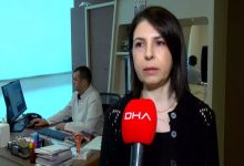 Bir sağlık merkezinde röportaj veren bir kadın, elinde kırmızı mikrofonla kameraya konuşuyor. Arka planda bir doktor bilgisayar başında çalışıyor; masa üzerinde tıbbi materyaller ve bir rahim modeli bulunuyor. Ortam klinik ve düzenli görünüyor.