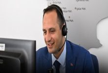 Ofis ortamında, takım elbiseli bir adam kulaklıkla bilgisayar ekranına bakarak gülümsüyor. Bir çağrı merkezi ya da destek hattında çalıştığı izlenimi veriyor; arka planda ise harita benzeri bir pano bulunuyor.