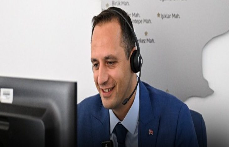 Ofis ortamında, takım elbiseli bir adam kulaklıkla bilgisayar ekranına bakarak gülümsüyor. Bir çağrı merkezi ya da destek hattında çalıştığı izlenimi veriyor; arka planda ise harita benzeri bir pano bulunuyor.