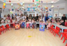 Bir anaokulu sınıfında çok sayıda çocuk ve öğretmen toplu fotoğraf çektiriyor. Sınıf renkli süslemeler, küçük sandalyeler ve oyuncaklarla dolu. Ön tarafta ağız ve diş sağlığına dikkat çeken afişler bulunuyor; ortam bir eğitim etkinliğini yansıtıyor.