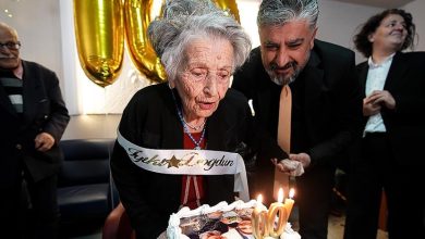 Yaşlı bir kadın, üzerinde “İyi ki doğdun” yazılı kuşakla, üzerinde fotoğraf bulunan doğum günü pastasındaki “100” mumlarını üflerken görülüyor. Yanında bir erkek ona destek olurken, arka planda altın renkli “100” balonlar ve kutlamaya katılan kişiler yer alıyor. Atmosfer sıcak ve duygusal bir 100. yaş kutlamasını yansıtıyor.