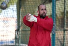 Kırmızı eşofmanlı bir sporcu, çekiç atma kafesinde atışa hazırlanırken görülüyor. Eldivenli elleriyle çekiç topunu savururken odaklanmış bir ifadeye sahip. Görseldeki kişi, işitme engelli milli atlet Muhammed Çakır.