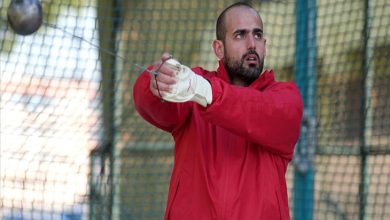 Kırmızı eşofmanlı bir sporcu, çekiç atma kafesinde atışa hazırlanırken görülüyor. Eldivenli elleriyle çekiç topunu savururken odaklanmış bir ifadeye sahip. Görseldeki kişi, işitme engelli milli atlet Muhammed Çakır.