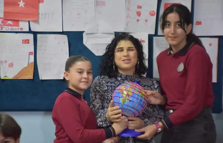 Fotoğrafta bir sınıf ortamında ortada oturan bir öğretmen ile yanında iki öğrenci görülüyor. Öğrenciler öğretmenle birlikte renkli bir dünya küresi tutuyor. Arka planda duvara asılmış öğrenci çizimleri ve Türk bayrağı bulunan sınıf panosu yer alıyor.