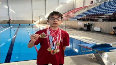 Kapalı bir yüzme havuzunda çekilmiş fotoğrafta, gözlüklü bir çocuk sporcu havuz kenarında ayakta duruyor. Boynunda çok sayıda madalya asılı, elinde ise bir madalyayı kameraya doğru tutuyor. Kırmızı tişört ve yüzme şortu giymiş. Arka planda havuz, kulvarlar ve tribün koltukları görülüyor; sahne net biçimde sportif bir başarı anını yansıtıyor.