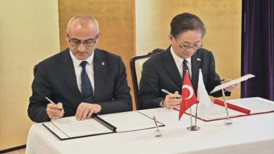 Bir masada yan yana oturan iki resmi giyimli kişi, önlerindeki belgeleri imzalıyor. Masanın üzerinde Türk bayrağı ve Japon bayrağı küçük masa bayrakları olarak yer alıyor. Ortam, resmi bir anlaşma veya protokol imza törenini andırıyor.