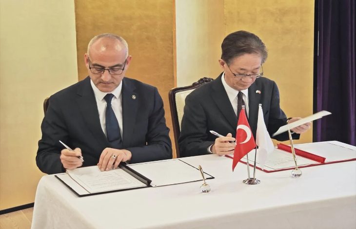 Bir masada yan yana oturan iki resmi giyimli kişi, önlerindeki belgeleri imzalıyor. Masanın üzerinde Türk bayrağı ve Japon bayrağı küçük masa bayrakları olarak yer alıyor. Ortam, resmi bir anlaşma veya protokol imza törenini andırıyor.