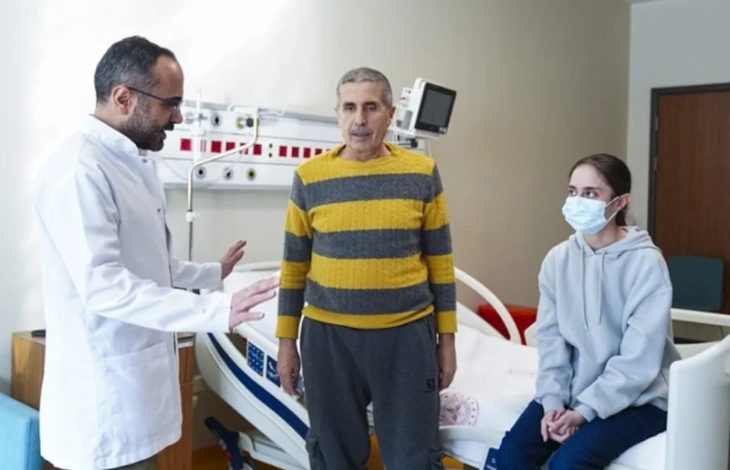 Fotoğrafta bir hastane odasında doktor, hasta ve yanında bulunan genç bir kadın görülüyor. Hasta ayakta dururken doktor onunla konuşuyor, genç kadın ise yatak kenarında oturarak durumu dikkatle izliyor. Ortam, organ nakli süreciyle ilgili bir hastane kontrolünü yansıtıyor.