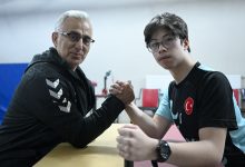 Fotoğrafta bir masa başında karşılıklı oturan iki kişi görülüyor. Biri orta yaşlı, diğeri genç olan iki kişi ellerini kenetleyerek bilek güreşi pozisyonu almış. Genç olan kişi Türkiye bayrağı bulunan spor tişörtü ve gözlük takıyor. İkisi de kameraya bakarken spor ve antrenman ortamını andıran bir mekânda bulunuyor.