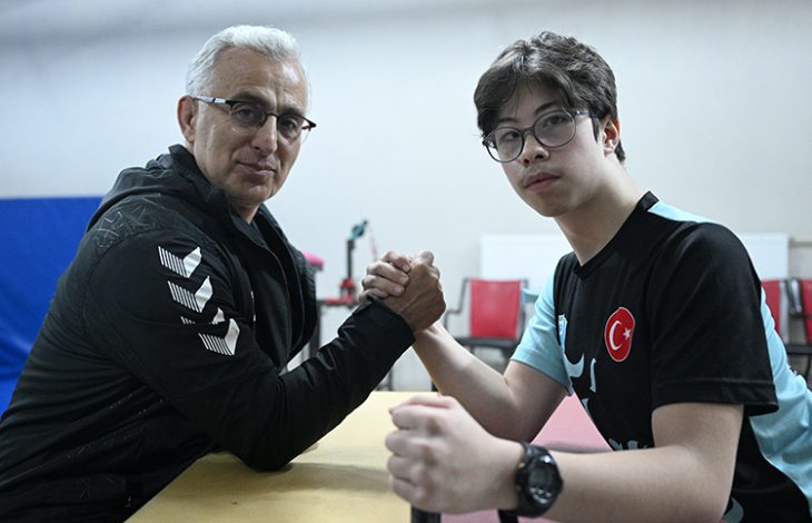 Fotoğrafta bir masa başında karşılıklı oturan iki kişi görülüyor. Biri orta yaşlı, diğeri genç olan iki kişi ellerini kenetleyerek bilek güreşi pozisyonu almış. Genç olan kişi Türkiye bayrağı bulunan spor tişörtü ve gözlük takıyor. İkisi de kameraya bakarken spor ve antrenman ortamını andıran bir mekânda bulunuyor.