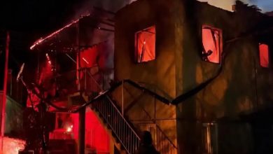 Gece saatlerinde iki katlı bir evin alevler içinde kaldığı görülüyor. Pencerelerden yoğun turuncu ışık yayılırken çatının bir kısmı çökmüş, merdiven ve dış cephe is içinde. Ön planda bir kişi yangını izlerken ortamda yoğun duman ve acil durum hissi hakim.