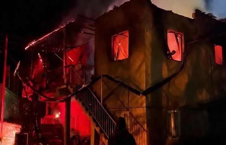 Gece saatlerinde iki katlı bir evin alevler içinde kaldığı görülüyor. Pencerelerden yoğun turuncu ışık yayılırken çatının bir kısmı çökmüş, merdiven ve dış cephe is içinde. Ön planda bir kişi yangını izlerken ortamda yoğun duman ve acil durum hissi hakim.