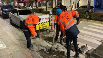 Gece saatlerinde şehir merkezinde, turuncu reflektif kıyafetli belediye ekipleri kaldırımda çalışma yapıyor. Üç görevli kazı yaparken yanlarında “Dikkat çalışma var” uyarı levhası bulunuyor. Arka planda park halindeki araçlar ve mağazalar görülüyor; sahne, erişim düzenlemesi ya da yol iyileştirme çalışmasını andırıyor.