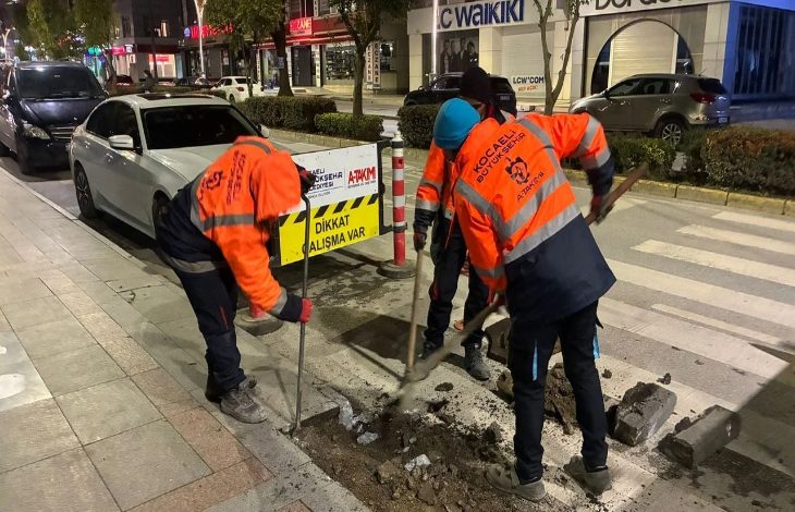 Gece saatlerinde şehir merkezinde, turuncu reflektif kıyafetli belediye ekipleri kaldırımda çalışma yapıyor. Üç görevli kazı yaparken yanlarında “Dikkat çalışma var” uyarı levhası bulunuyor. Arka planda park halindeki araçlar ve mağazalar görülüyor; sahne, erişim düzenlemesi ya da yol iyileştirme çalışmasını andırıyor.