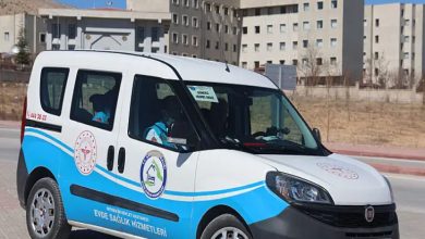 Görselde, bir hastanenin önünde park etmiş “Evde Sağlık Hizmetleri”ne ait resmi bir hizmet aracı görülüyor. Üzerinde sağlık birimlerinin amblemleri ve iletişim bilgileri yer alan araç, açık havada gündüz saatlerinde çekilmiş. Arka planda büyük bir eğitim ve araştırma hastanesi binası bulunuyor.