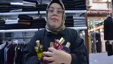 Fotoğrafta başörtülü ve gözlüklü genç bir kadın, bir giyim mağazasının içinde kollarında birkaç parfüm şişesi tutarak kameraya doğru duruyor. Arka planda raflara dizilmiş pantolonlar ve askıda kıyafetler görülüyor; mağaza düzenli ve aydınlık bir çarşı dükkânı atmosferi veriyor.