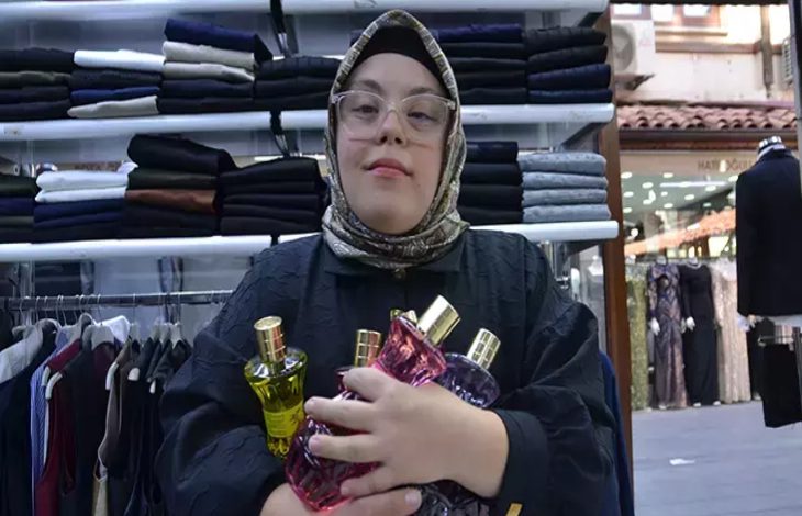 Fotoğrafta başörtülü ve gözlüklü genç bir kadın, bir giyim mağazasının içinde kollarında birkaç parfüm şişesi tutarak kameraya doğru duruyor. Arka planda raflara dizilmiş pantolonlar ve askıda kıyafetler görülüyor; mağaza düzenli ve aydınlık bir çarşı dükkânı atmosferi veriyor.