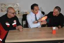 Bir masa etrafında üç erkek oturuyor. Ortadaki kişi, yanındaki adamın koluna tuttuğu cihazla damarları görünür hale getiriyor; cihazın yaydığı ışıkla kol üzerinde damarlar belirgin şekilde parlıyor. Soldaki kişi ise dikkatle süreci izliyor. Ortam, bir eğitim ya da tanıtım anını andırıyor.