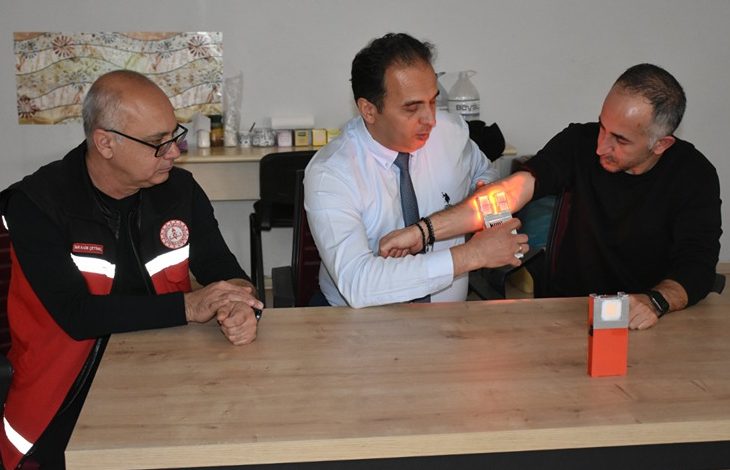 Bir masa etrafında üç erkek oturuyor. Ortadaki kişi, yanındaki adamın koluna tuttuğu cihazla damarları görünür hale getiriyor; cihazın yaydığı ışıkla kol üzerinde damarlar belirgin şekilde parlıyor. Soldaki kişi ise dikkatle süreci izliyor. Ortam, bir eğitim ya da tanıtım anını andırıyor.